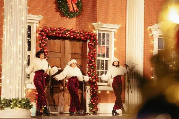 Three female singers perform festive holiday music on the courthouse steps at Merry & Bright at Martini Town, surrounded by Christmas lights and decorations.